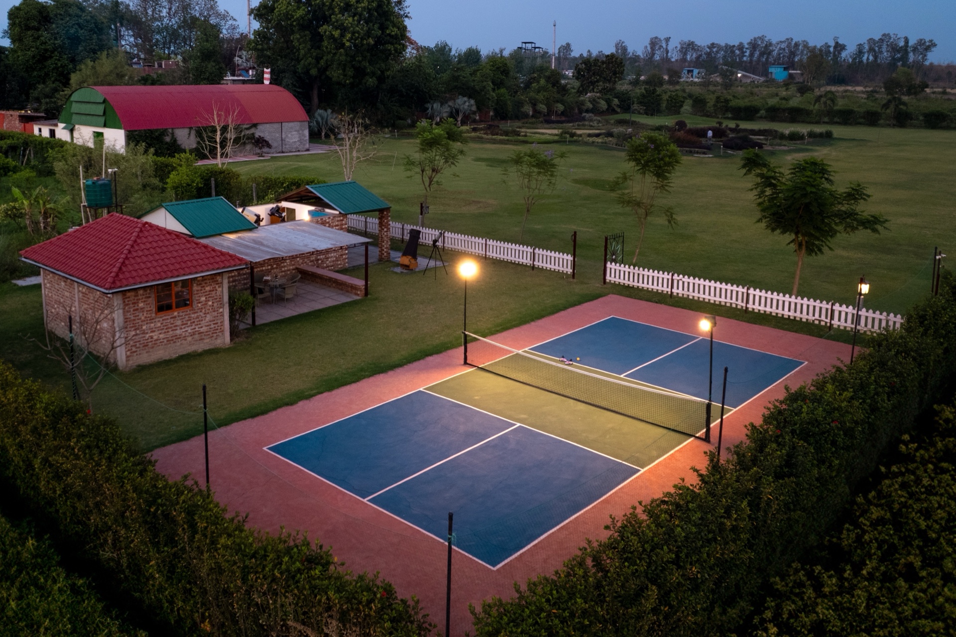 The farm's pickleball court lit at dusk, beside the lawns and the red-roofed hangar